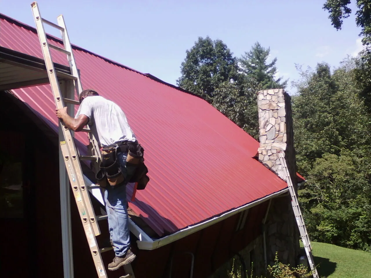 Roofer on a ladder beside a red metal roof and stone chimney — Soffit & Fascia Repair by Roofing Brothers in Rock Spring